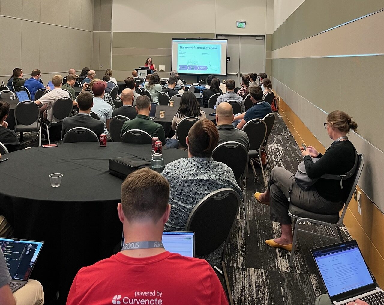 Image of a room of round tables with black covers filled with people listening to my talk. At the front is a speaker wearing a red tank top. On the screen is a slide that says 'The Power of Community Review.'' style='width: 90%;