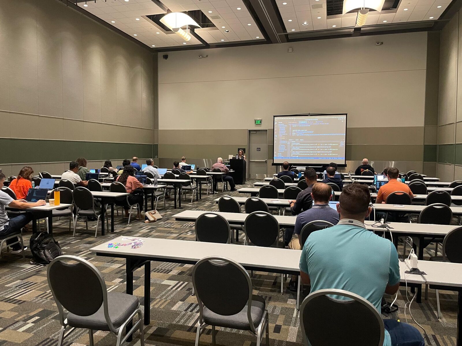 A photograph showing a large open and well-lit meeting room with two rows of horizontal tables. People are working at laptops in the room. An instructor is at the front with their laptop screen projected on the screen' style='width: 90%;