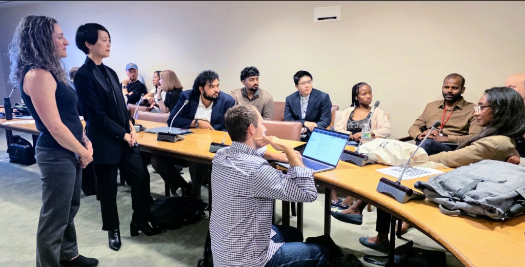 Maintainers gather around a conference table during a Maintain-a-Thon breakout session at UN Open Source Week, with two facilitators standing at the head of the table.